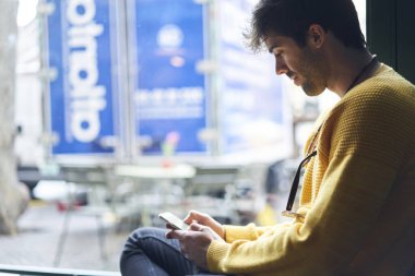 Sıradan giyinen yakışıklı bir erkeğin pencere eşiğinde şehir manzaralı büyük bir pencereye karşı otururken cep telefonu takmasının yan görüntüsü