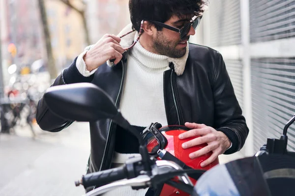 Content bearded ethnic male in casual outfit and sunglasses looking away while resting on scooter after ride in sunny day