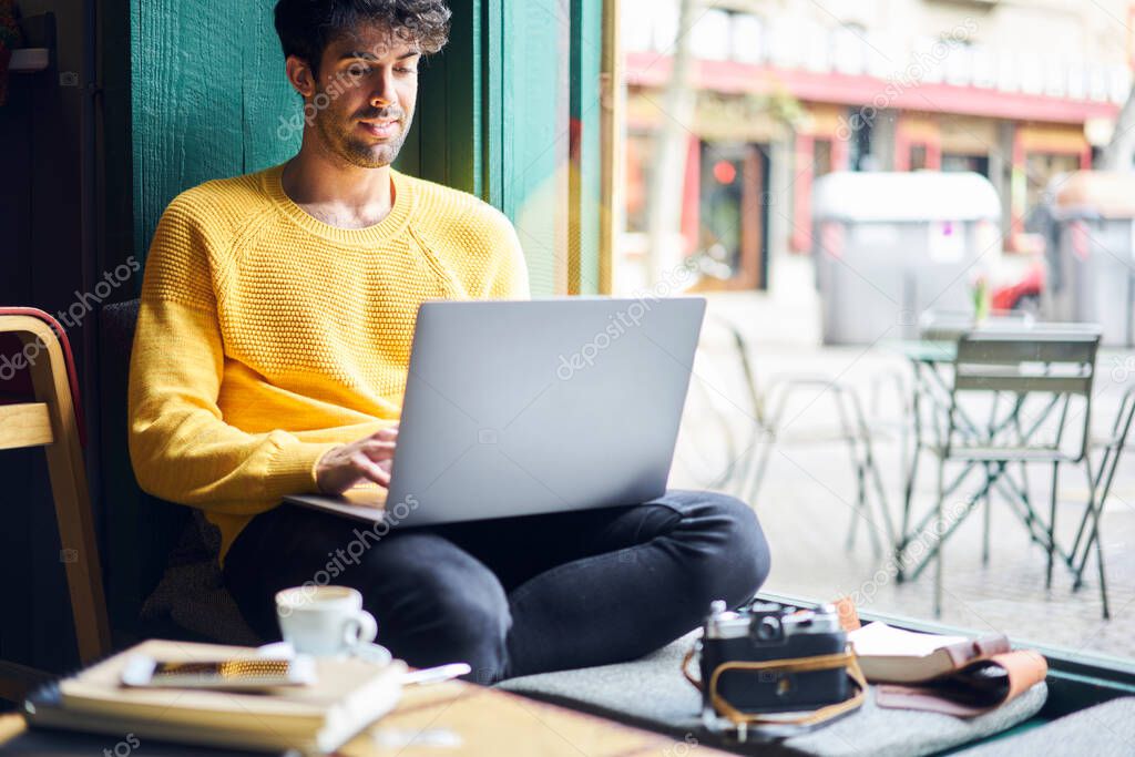Pensive ethnic male freelancer in casual wear sitting at wooden table in cozy cafe and working on remote project on laptop