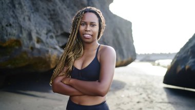 Genç Afro-Amerikan sportif ve formda bayan koç kameraya gülümseyerek bakıyor ve açık hava antrenmanı sırasında kayalık sahil şeridinde duruyor.