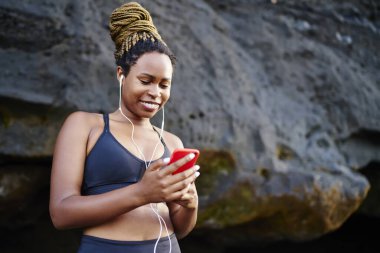 Genç Afro-Amerikan formda koşucu gülümseyen ve kayalık sahilde duran eğitim sırasında akıllı telefona bakarken yere bakan.