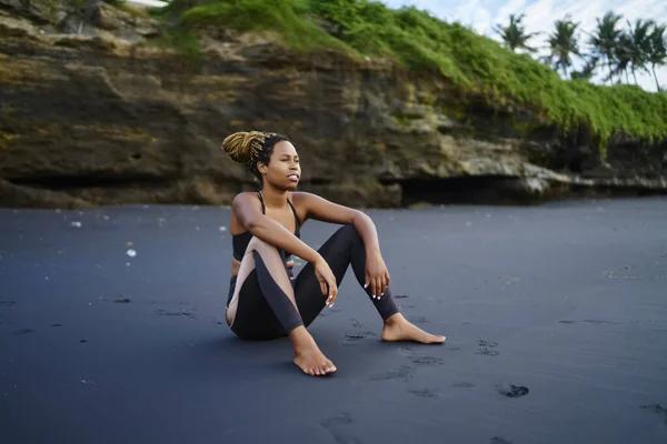 Full length of exhausted barefoot African American female in sportswear looking away while sitting on beach with black sand resting after workout