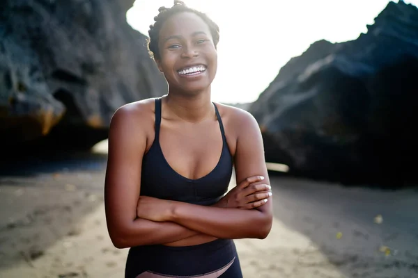 Content African American female in sportswear looking at camera and smiling while resting on sandy beach after training in summer