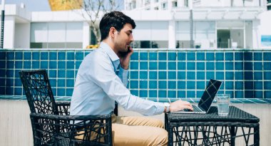 Günışığında modern şehrin terasında çalışan, netbook 'ta yazan, akıllı telefondan konuşan, resmi tişörtlü etnik adamın yan görünüşü