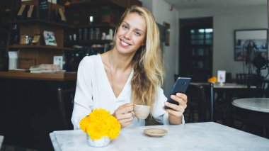 Çekici, güler yüzlü bir bayan, elinde bir fincan lezzetli kahveyle küçük bir vazoda canlı sarı çiçeklerle oturmuş kameraya bakıyor ve çağdaş kafede akıllı telefon kullanıyor.