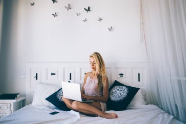 Ciddi sarışın, ipek pijamalı bayan öğrenci güneşli bir günde Barcelona 'da rahat bir yatakta dizüstü bilgisayarla bilgi arıyor.