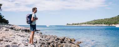 Güneşli bir günde bulutlu gökyüzüne karşı mavi denizin taşlı sahilinde duran sırt çantalı yetişkin adamın yan görüntüsü
