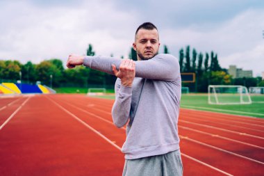 Gerçekten formda sakallı genç bir antrenör kollarını esnetiyor. Stadyumda hava bulutluyken sıkı çalıştıktan sonra dinleniyor.