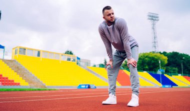 Ciddi sakallı, eşofmanlı genç bir koşucunun stadyumdaki sıkı antrenmandan sonra ara verip bakışlarını kaçırmasından.