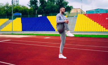 Genç güçlü erkek sporcu ısınmak için egzersiz yapıyor sabah stadyumda ayakta antrenman yaparken gri eşofmanlı 20 'li yaşlarda beyaz sporcu spor yaparken spor yapma hedeflerine ulaşıyor.