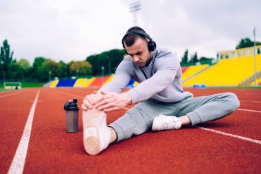 Spor kıyafetli güçlü bir sporcu kauçuk pistte oturur ve müzik dinlerken bacak egzersizi yapar.