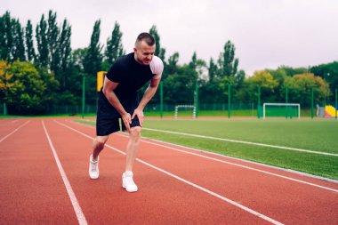 Stadyumda antrenman yaparken dizinden sakatlanan şortlu ve tişörtlü üzgün, profesyonel bir erkek koşucunun tüm vücudu.