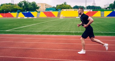Kulaklıklı, aktif giyinmiş, stadyumda koşan genç, yakışıklı bir erkek spor hedeflerine ulaşıyor. Güçlü bir sporcu, sabah kardiyo antrenmanına hazır ve kulaklıklı favori müzikle çıkıyor.