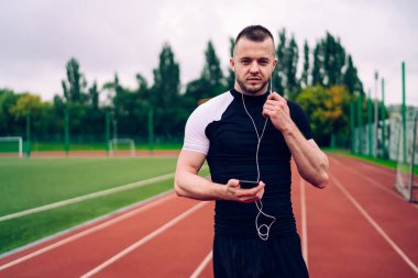 Forma girmiş genç kaslı erkek atlet akıllı telefondan müzik dinliyor bulutlu bir yaz gününde stadyumda koşan kameraya bakıyor.