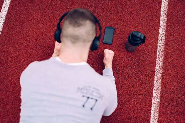 From above back view of anonymous young guy with phone and water thermos listening to song in headphones while doing plank workout on stadium track