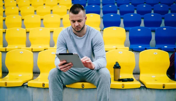 Serious male runner recording his results for training routine using digital tablet and 4G connection, pensive man in tracksuit sending mails and searching information about workout and exercises