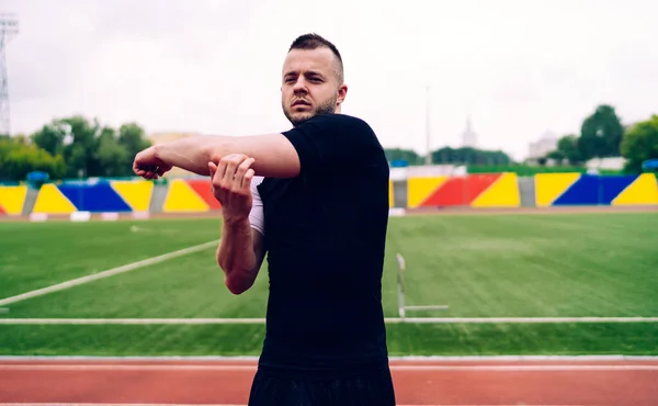 Serious caucasian male jogger doing warming up exercises for upper body prepare for running on stadium, determined hipster guy keeping healthy lifestyle slimming and losing weight for wellness