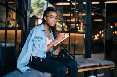 Güzel Afro-Amerikan hippi kız gözlüklü portresi elinde not defteri ve kafenin içinde boş zaman üzerine yazı yazan modaya uygun giyinmiş genç bir kadın kameraya bakıyor ve boş zamanlardaki hobi üzerine çizimler yapıyor.