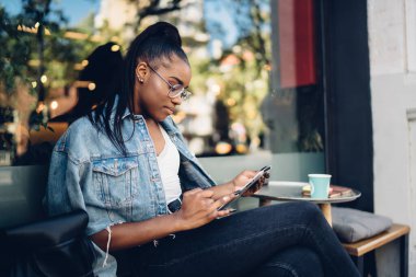 Afro-Amerikalı genç bir bayanın sosyal medyada tabletle mesajlaşması ve bir fincan kahveyle sokak kafesinde dinlenmesi.