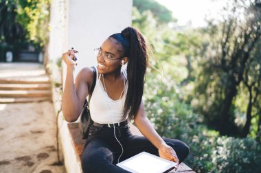 Uzun saçlı, yaz kıyafetleri ve gözlüklü bir kadın güneşli bir günde parktaki korkulukta müzik dinlerken gözlük takıyor ve başka tarafa bakıyordu.