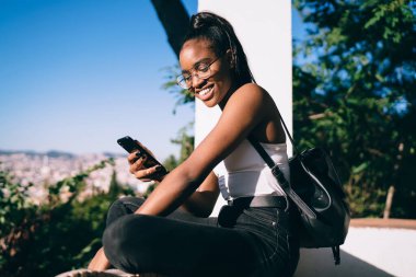 Sırt çantalı, sosyal medyada akıllı telefondan sohbet eden ve gündüz vakti balkonda gülümseyen neşeli Afro-Amerikan kadın.