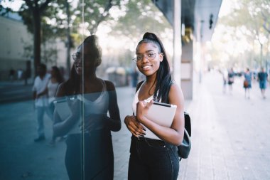 Modern binanın penceresinin yanında gündelik giysiler içinde duran ve gündüz vakti mola verirken cam duvara yansıyan pozitif Afrikalı genç bir kadın.
