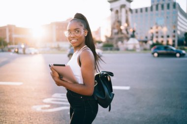 Şehre yakın bir yerde sırt çantalı ve tableti incelerken kameraya gülümseyen genç bir etnik kadın.