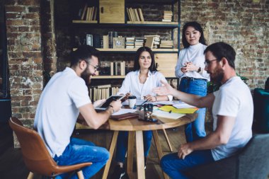 Bir grup çok ırklı iş arkadaşı, günlük kıyafetlerini giymiş, ahşap yuvarlak masa başında oturmuş, çalışma stratejilerini tartışıyorlar.