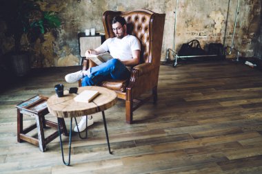 Yüksek açılı, uzun sakallı, beyaz gömlekli, gözlüklü, deri koltukta oturan ve ofisteki kitaptan keyif alan bir erkek.