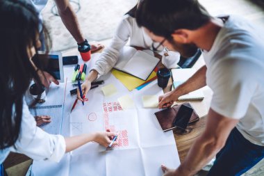 Erkek ve bayan meslektaşlar tarafından çevrelenmiş masanın üstünden proje çizelgeleri ve istatistikleri analiz ederek iş stratejisi üzerinde birlikte çalışırlar.