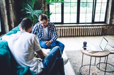 Erkek iş arkadaşlarının dokümanlarla birlikte çalıştığı yüksek açı, dizüstü bilgisayarlı ve kahveli koltuğun yanında otururken,