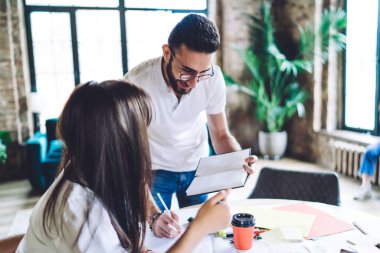 Modern gözlüklü ve gündelik kıyafetli bir adam modern odadaki bayan meslektaşıyla proje tartışırken kağıda not alıyor.