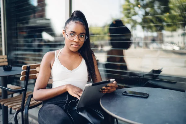 穿着休闲装 头脑清醒的非洲裔美国女人坐在桌旁 一边用智能手机 一边在平板电脑上冲浪 一边在街上的咖啡店休息 — 图库照片