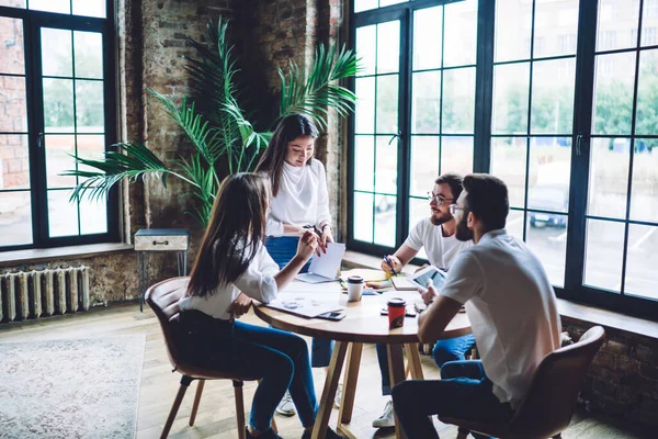 Content Asian female manager in casual outfit standing in creative workplace and explaining business strategy to colleagues sitting at wooden table