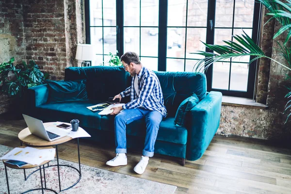 Full body of businessman in casual clothes sitting on sofa near table with smartphone coffee and laptop while looking through documents at workplace