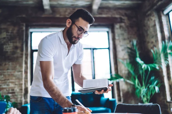 From below of serious bearded male entrepreneur wearing stylish eyeglasses and casual clothes writing notes and looking down with big window on background