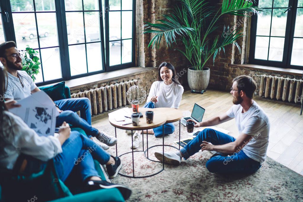 Female and male entrepreneurs in casual clothes sitting on floor and sofa while brainstorming and discussing business ideas in cozy apartment
