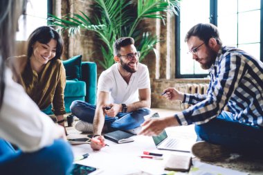 Yaratıcı çalışma alanında çalışırken ve yerde otururken gülümserken gündelik kıyafet çizimi projesinde çalışan çok ırklı meslektaşlar.