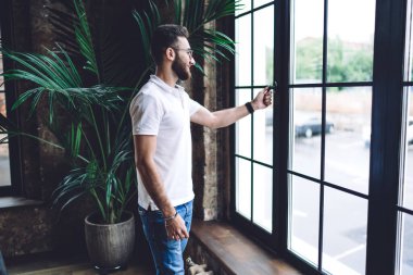 Gündüz vakti modern ofiste günlük giysiler ve gözlüklerle ayakta duran ve pencereyi açan pozitif sakallı etnik adamın yan görünüşü