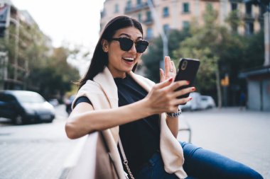 Güneş gözlüklü ve günlük giyimli mutlu Asyalı kadın gezen, neşeli blog yazarı ile iletişim kurmak için akıllı telefonu kullanan cep telefonu kullanıyor.