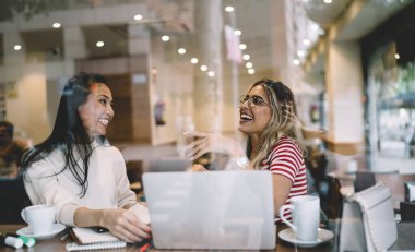 Gülümseyen milenyum yeniyetme kız öğrencileri modern dizüstü bilgisayar kullanarak online kurslar hakkında konuşuyorlar, neşeli genç kadınlar kafeteryada buluşurken ve iletişim kurarken fikirlerini paylaşıyorlar.