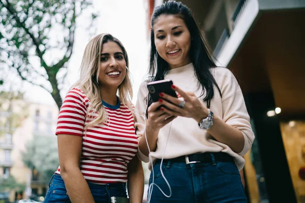 カメラで笑顔スペイン千年紀の女の子の半分の長さの肖像画一方 アジアの親友は一緒に娯楽中に聞くためのオーディオセットを選択しながら 幸せな観光客は 携帯電話上のオンラインガイドを追跡 — ストック写真