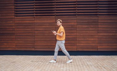 Sokakta yürürken ahşap duvara yaslanmak için elinde kahveyle gezen aktif stil sahibi genç adamın yan görüntüsü