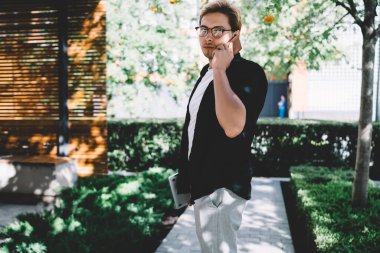 Moda gözlüklü, siyah gömlekli, elinde laptop ve cep telefonuyla şehir meydanında dikilen şık ve şık bir gencin yan görüntüsü