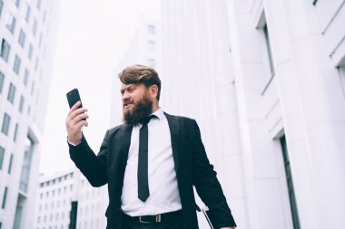 Şehir caddesinde dikilip hoşnutsuzca akıllı telefon ekranına bakan akıllı takım elbiseli çok kızgın bir girişimci.