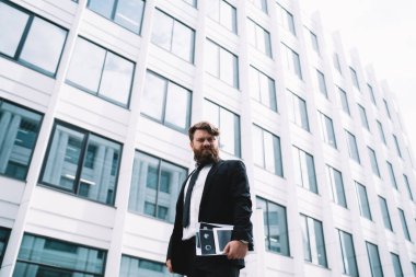 Kendine güvenen sakallı iş adamının resmi giysileri ve kravatıyla elinde belgelerle modern binanın yanında durduğu düşük açı.