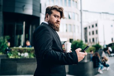 Resmi görevdeki düşünceli sakallı işadamının yan görünüşü caddede dikilip akıllı telefonlar kullanarak kameraya bakarken bir fincan kahve içmesi.