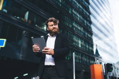 Resmi takım elbiseli, kendine güvenen, tablet giyen, arka planda modern yüksek kulesi olan ve başka yöne bakan bir adamdan.