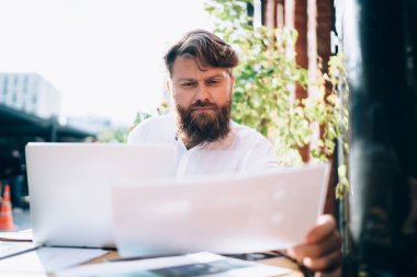 Yoğunlaşmış sakallı, beyaz gömlekli, serbest çalışan, açık kafedeki ahşap masada oturan ve dizüstü bilgisayarını incelerken kağıtlarla çalışan bir erkek.