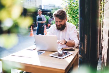 Yoğunlaştırılmış sakallı, uzaktan kumandalı bir erkek, dışarıda, tablet ve akıllı telefonlu bir kafede çalışırken dizüstü bilgisayarda yazıyor.